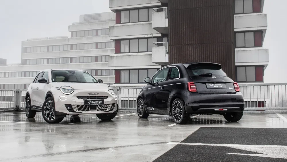 Een witte Fiat 600 en een zwarte Fiat 500 op een nat parkeerdek met mistige flats op de achtergrond.